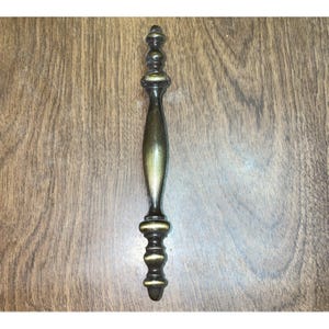 May include: A vertical metal cabinet pull handle featuring an antique brass finish with a dark patina. The handle has a central elongated spindle shape flanked by decorative tiered, rounded finials at both the top and bottom ends.