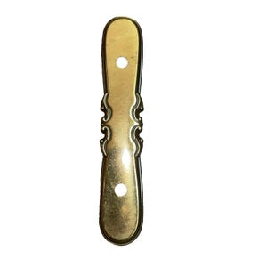 May include: A gold-coloured metal furniture pull with two holes for mounting. The pull has an elongated shape with decorative scrollwork in the centre. The edges are dark, giving it a vintage appearance.