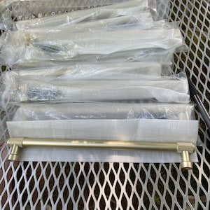 May include: A brushed gold cabinet pull with a cylindrical bar and textured, knurled ends. The handle is presented horizontally, resting on a white foam protective sleeve, with several additional identical pulls packaged in clear plastic bags behind it.