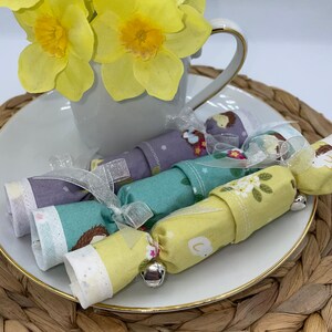 May include: Fabric crackers in pastel shades, patterned with floral and animal motifs, tied with ribbons and small bells. They sit on a white plate with a gold rim, next to yellow daffodils in a white teacup, creating a spring theme.