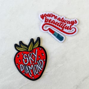 May include: Two embroidered iron-on patches. One patch is red and white and says "You're always beautiful". The other patch is red and green and says "Sky Diamond" with a strawberry design.