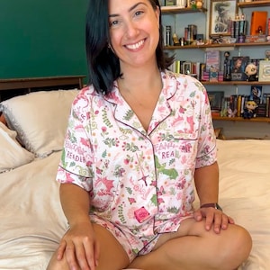 Puede incluir: Conjunto de pijama rosa con tem&aacute;tica de fantas&iacute;a. La camiseta de manga corta y los pantalones cortos est&aacute;n cubiertos con un estampado de dragones, setas y elementos florales. Las palabras "Fantasy Reader" est&aacute;n impresas en la parte superior. El conjunto tiene ribetes rosa oscuro.