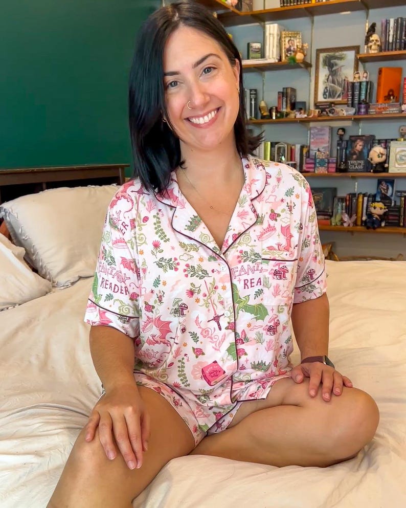 Puede incluir: Conjunto de pijama rosa con tem&aacute;tica de fantas&iacute;a. La camiseta de manga corta y los pantalones cortos est&aacute;n cubiertos con un estampado de dragones, setas y elementos florales. Las palabras "Fantasy Reader" est&aacute;n impresas en la parte superior. El conjunto tiene ribetes rosa oscuro.