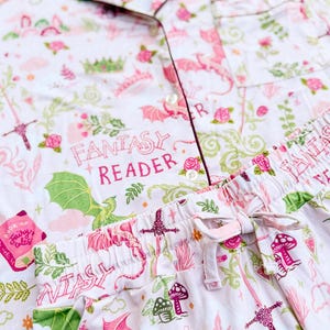 Puede incluir: Conjunto de pijama blanco con tem&aacute;tica de fantas&iacute;a rosa. El conjunto presenta una camisa con botones y pantalones con cord&oacute;n, adornados con dragones, espadas, setas y el texto "FANTASY READER". El ribete es de color rosa oscuro.