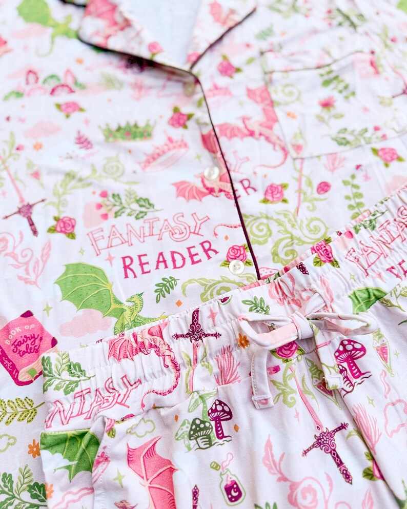 Puede incluir: Conjunto de pijama blanco con tem&aacute;tica de fantas&iacute;a rosa. El conjunto presenta una camisa con botones y pantalones con cord&oacute;n, adornados con dragones, espadas, setas y el texto "FANTASY READER". El ribete es de color rosa oscuro.