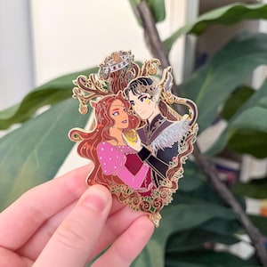May include: A gold enamel pin with a romantic illustration of two figures. The female figure has long red hair and wears a pink and red dress. The male figure has black hair and wears a black coat. The pin has a decorative border.
