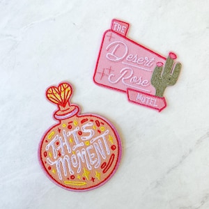 May include: Two embroidered iron-on patches. One is pink and says "The Desert Rose Motel" with a green cactus. The other is peach and says "This Moment" with a heart and a bottle.