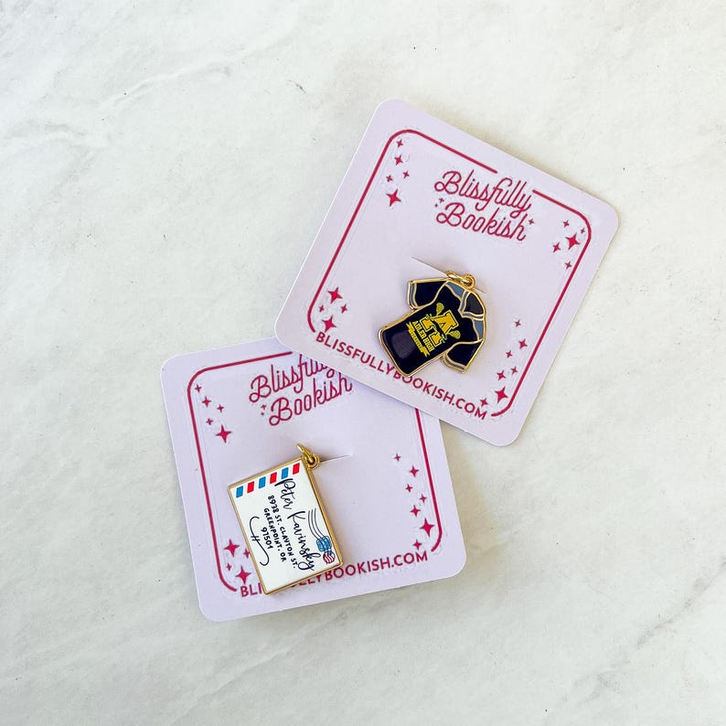 May include: Two enamel pins featuring a black t-shirt with a gold design and a letter with a red, white, and blue design. The pins are on white cards with pink stars and the text "Blissfully Bookish" and "blissfullybookish.com".