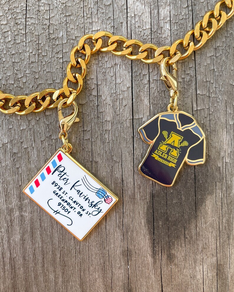 May include: Gold chain with two charms. One charm is a black and gold lacrosse jersey with the text "Adler High" and the other charm is a white envelope with blue and red stripes and the text "Peter Kavinsky 8938 St. Clayton St. Greenpoint, OR 97501".