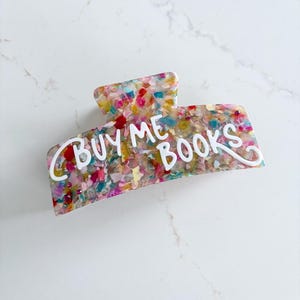 May include: A colorful hair claw clip with a confetti-like design. The clip features the white text "BUY ME BOOKS" in a playful font. The clip is made of a shiny, durable material.