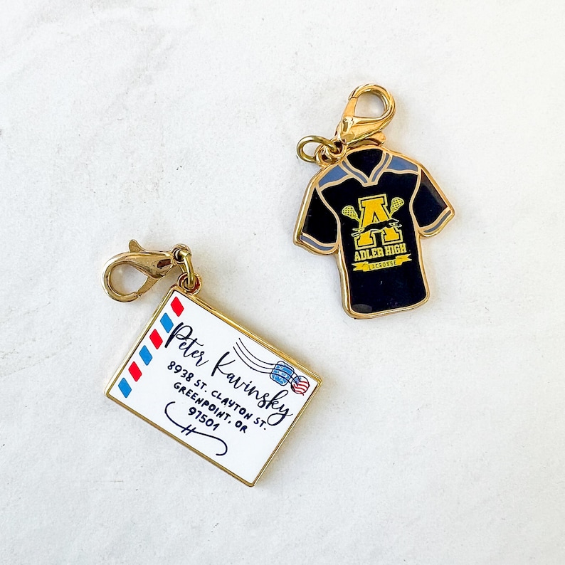 May include: Two gold-colored metal charms. One charm is a black lacrosse jersey with a gold letter A and the words "Adler High Lacrosse". The other charm is a gold envelope with the words "Peter Kavinsky 8928 St. Clayton St. Greenpoint, OR 97501" written on it.