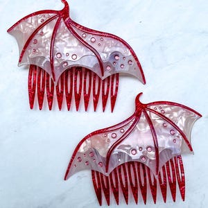 May include: Two red and white hair combs with a bat wing design. The combs have a red base with a wing-shaped top in translucent white and red, featuring circular accents. The combs are displayed on a white marble surface.