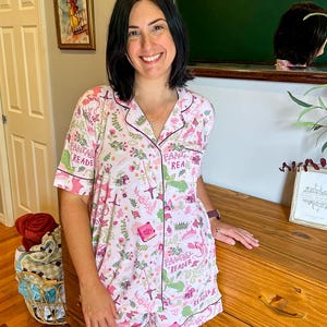 Puede incluir: Un conjunto de pijama rosa claro con un estampado floral y de fantas&iacute;a. El conjunto incluye una camisa con botones de manga corta y pantalones cortos a juego, ambos con ribetes de color rosa oscuro. Las palabras "Fantasy Reader" est&aacute;n impresas en el conjunto.