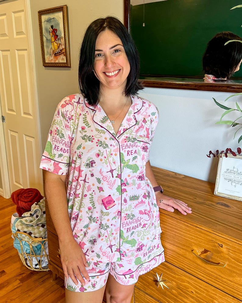 Puede incluir: Un conjunto de pijama rosa claro con un estampado floral y de fantas&iacute;a. El conjunto incluye una camisa con botones de manga corta y pantalones cortos a juego, ambos con ribetes de color rosa oscuro. Las palabras "Fantasy Reader" est&aacute;n impresas en el conjunto.