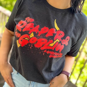May include: Dark gray t-shirt with the text "The Reaper of the Goblin King" in red and yellow. The graphic has a distressed, vintage look. The shirt is short-sleeved and appears to be made of a soft material.