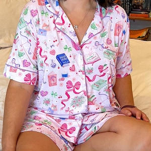 May include: A light pink pajama set featuring a pattern of bows, hearts, books, and the text "Fated Mates" and "Slow Burn". The short-sleeved top and shorts have pink edging.