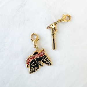 May include: Two gold-colored metal charms. One charm features a black hand with the words "DIRTY HANDS" in red on a banner. The other charm is a black stick with a white bird head on top.