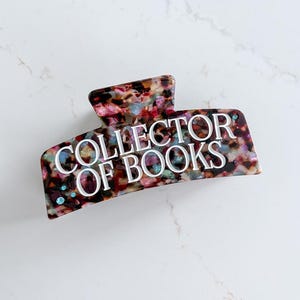 May include: A colorful hair claw clip with the text "COLLECTOR OF BOOKS" in white. The clip features a mix of colors including brown, pink, and blue. The clip is made of a shiny material and has a curved shape.