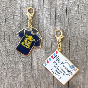 May include: Two gold-colored keychains. One keychain features a blue lacrosse jersey with the text "ADLER HIGH LACROSSE" and a gold letter "A". The other keychain features a white envelope with red and blue stripes and the text "Peter Kavinsky 8938 ST. CLAYTON ST. GREENPOINT, OR 97501".