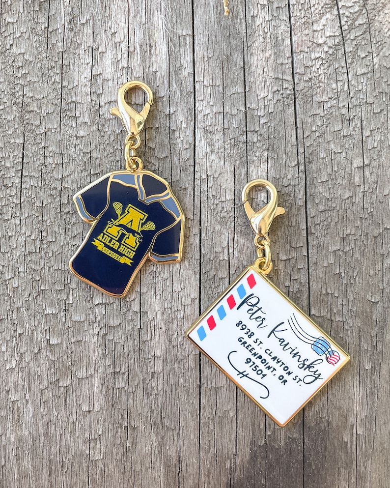 May include: Two gold-colored keychains. One keychain features a blue lacrosse jersey with the text "ADLER HIGH LACROSSE" and a gold letter "A". The other keychain features a white envelope with red and blue stripes and the text "Peter Kavinsky 8938 ST. CLAYTON ST. GREENPOINT, OR 97501".
