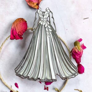 May include: A silver and white enamel pin in the shape of a wedding dress. The dress features a detailed design with a flowing skirt and floral accents on the bodice. The pin is surrounded by dried rose petals and a twine circle.