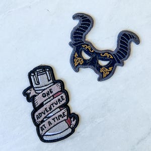 May include: Two embroidered iron-on patches. One patch is black with gold accents and features a masquerade mask with horns. The other patch is a light gray bottle with a black outline and the text "One Adventure at a Time".
