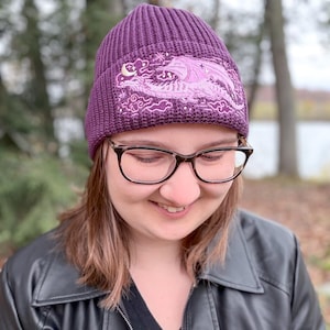 May include: A purple knit beanie with a detailed embroidered design featuring a dragon, moon, and stars. The beanie is a deep purple color and has a ribbed texture. The embroidered design is in shades of pink and purple.