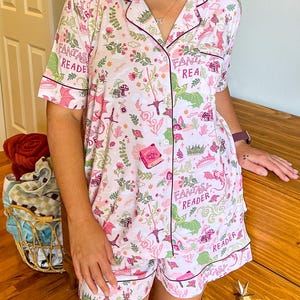 Puede incluir: Un conjunto de pijama rosa claro con tem&aacute;tica de fantas&iacute;a. El conjunto incluye una camisa de manga corta con botones y pantalones cortos, ambos adornados con un patr&oacute;n caprichoso de dragones, espadas, libros y el texto "Fantasy Reader". El ribete es rosa oscuro.