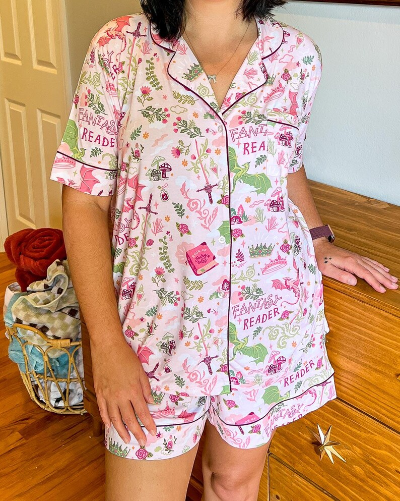 Puede incluir: Un conjunto de pijama rosa claro con tem&aacute;tica de fantas&iacute;a. El conjunto incluye una camisa de manga corta con botones y pantalones cortos, ambos adornados con un patr&oacute;n caprichoso de dragones, espadas, libros y el texto "Fantasy Reader". El ribete es rosa oscuro.