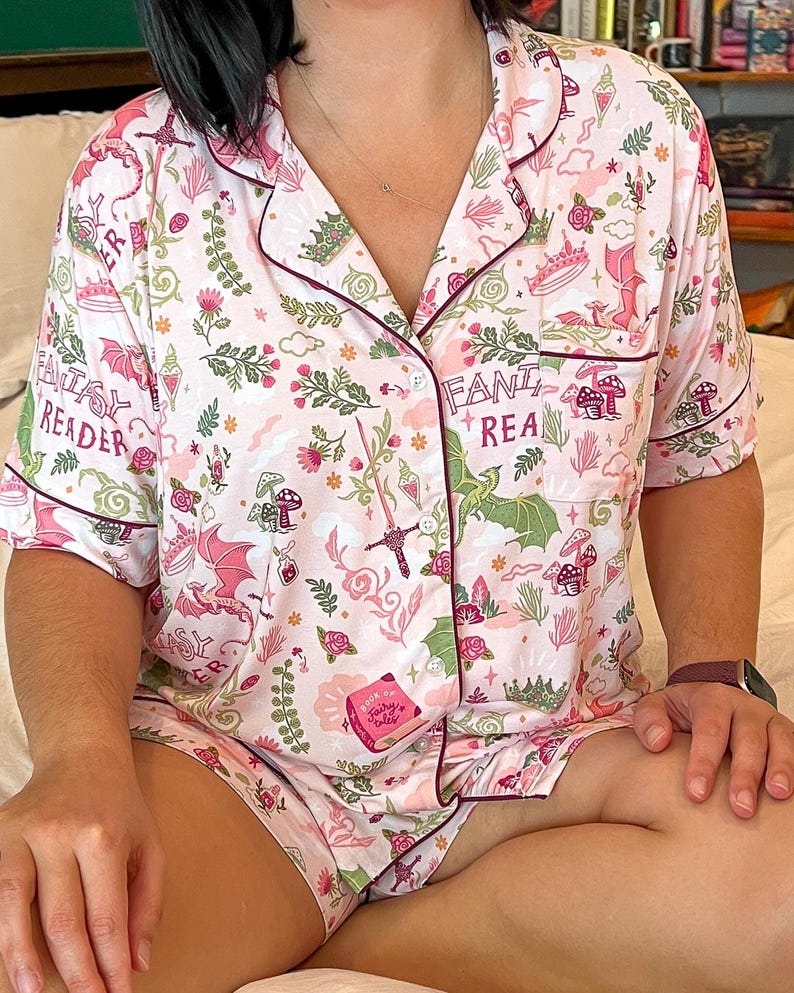 Puede incluir: Conjunto de pijama rosa con tem&aacute;tica de fantas&iacute;a. El conjunto incluye una camisa con botones y pantalones cortos, ambos adornados con un patr&oacute;n repetido de dragones, setas, espadas y las palabras "Fantasy Reader" y "Fantasy Realm". Ribete burdeos oscuro.