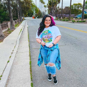 May include: A woman wearing a white t-shirt with the text "Fantasy Book Lover" in colorful letters and a blue plaid shirt tied around her waist. She is wearing blue jeans with rips and black sneakers.