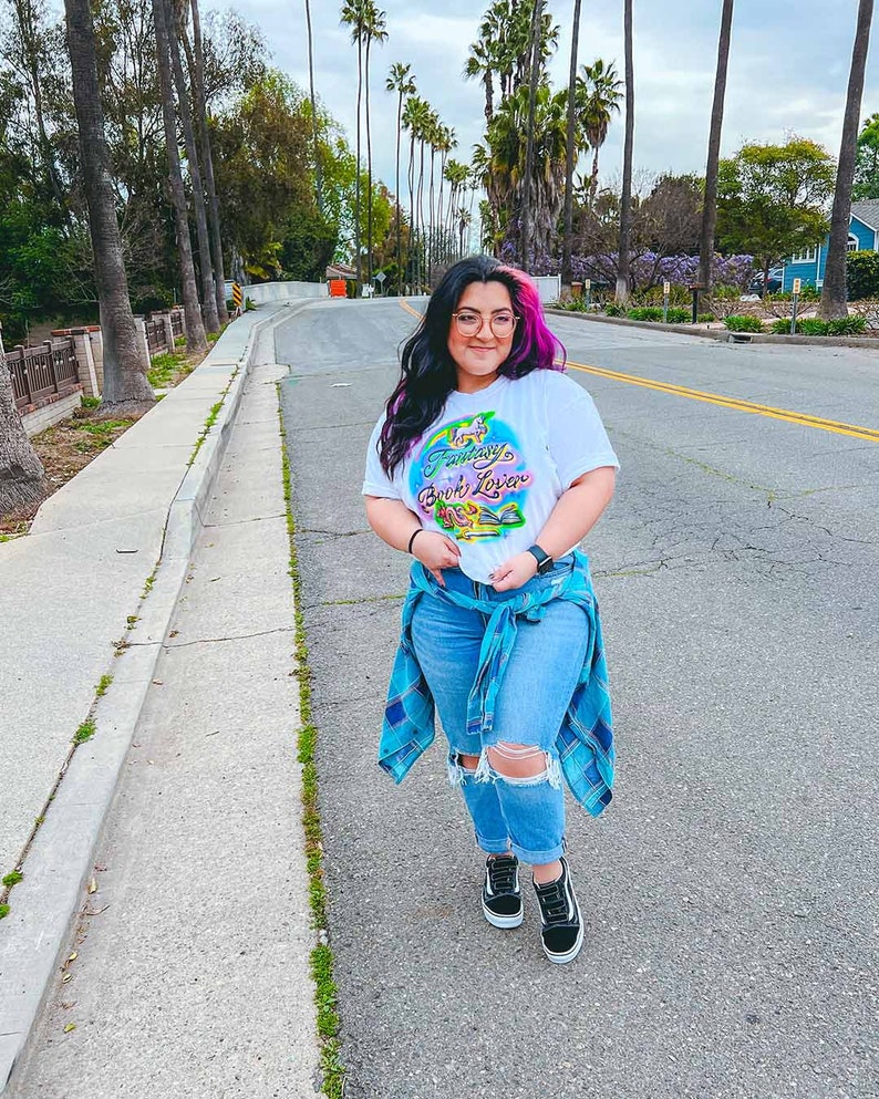 May include: A woman wearing a white t-shirt with the text "Fantasy Book Lover" in colorful letters and a blue plaid shirt tied around her waist. She is wearing blue jeans with rips and black sneakers.