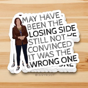 May include: A pile of white stickers featuring a quote and an illustration of a person. The text says "MAY HAVE BEEN THE LOSING SIDE STILL NOT CONVINCED IT WAS THE WRONG ONE." The illustration shows a person in a brown jacket, blue shirt, and dark trousers.