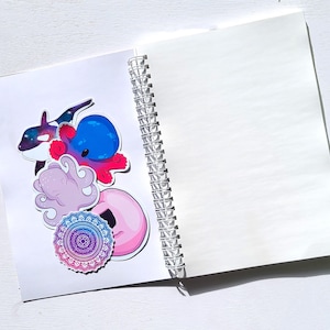 May include: A white spiral bound notebook with a variety of colorful stickers on the front cover. The stickers include an octopus, a whale, a mandala, and a donut.