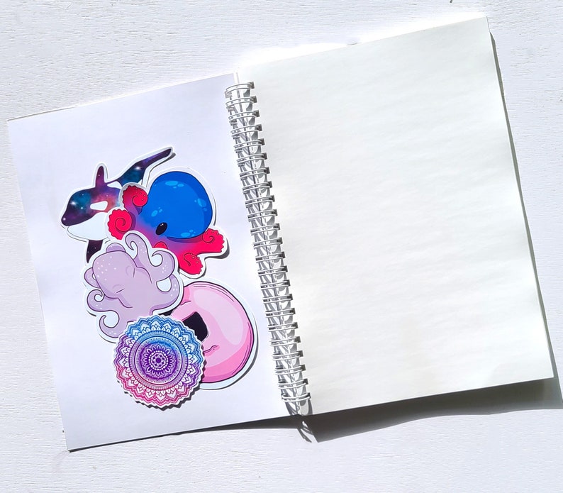 May include: A white spiral bound notebook with a variety of colorful stickers on the front cover. The stickers include an octopus, a whale, a mandala, and a donut.