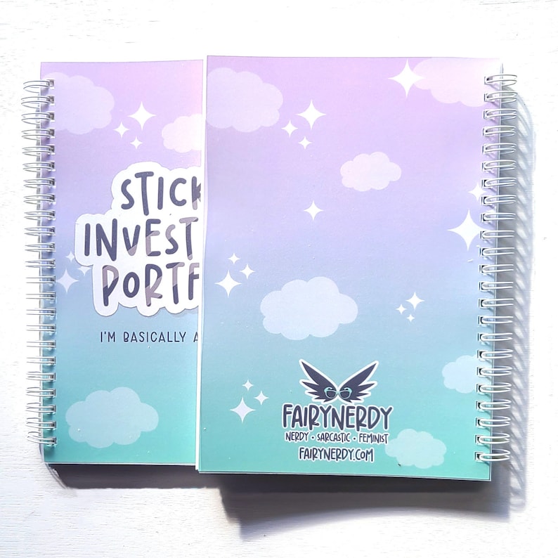 May include: A spiral bound notebook with a light blue and purple gradient background. The notebook has a sticker on the front that says "Stick to your investment portfolio, I'm basically a fairy godmother". The back of the notebook has a logo for Fairy Nerdy, which says "Nerdy. Sarcastic. Feminist. FairyNerdy.com".