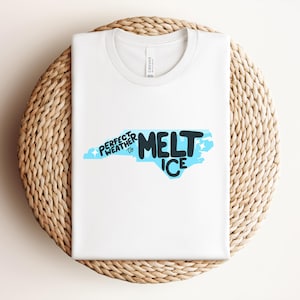 Puede incluir: Una camiseta blanca con el texto "Perfect Weather to Melt Ice" en un diseño azul y negro con la forma del estado de Carolina del Norte. La camiseta está doblada sobre una superficie redonda y tejida.