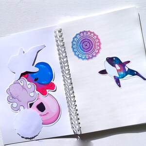 May include: A white spiral bound notebook with colorful stickers on the front cover. The stickers include a pink octopus, a blue and red octopus, a white dolphin, a donut, a white circle, and a purple and blue mandala.