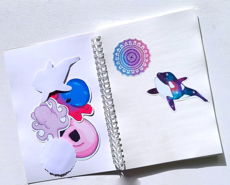 May include: A white spiral bound notebook with colorful stickers on the front cover. The stickers include a pink octopus, a blue and red octopus, a white dolphin, a donut, a white circle, and a purple and blue mandala.
