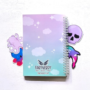 May include: A purple and blue notebook with a spiral binding. The notebook has a gradient background with clouds and stars. The cover features the text "FairyNerdy" and the website "fairynerdy.com". The notebook has three stickers on it: a purple octopus, a purple skull, and a pink and blue dolphin.