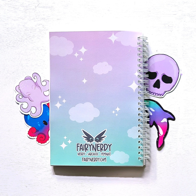 May include: A purple and blue notebook with a spiral binding. The notebook has a gradient background with clouds and stars. The cover features the text "FairyNerdy" and the website "fairynerdy.com". The notebook has three stickers on it: a purple octopus, a purple skull, and a pink and blue dolphin.