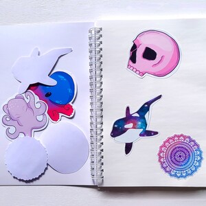 May include: An open notebook with a variety of stickers, including a pink skull, a blue and purple mandala, a purple octopus, a blue and white whale, and a white fish. The notebook has a spiral binding.