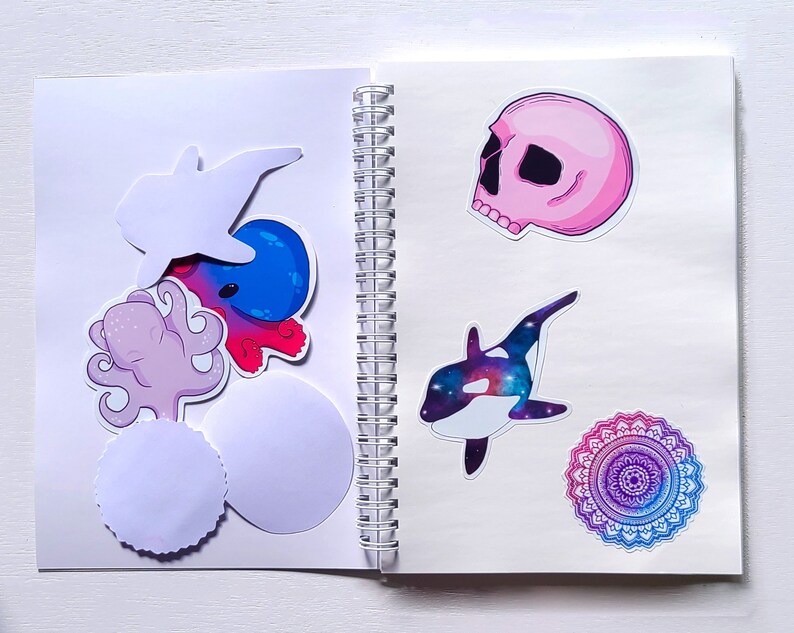 May include: An open notebook with a variety of stickers, including a pink skull, a blue and purple mandala, a purple octopus, a blue and white whale, and a white fish. The notebook has a spiral binding.