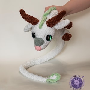 May include: A white and brown crochet dragon with large green eyes, brown horns, and a long, curved tail. The dragon is handmade and has a pink mouth and gray nose. The dragon is being held by a person.