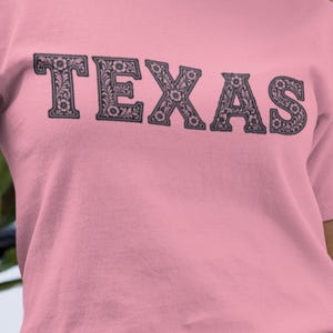 May include: A light pink t-shirt with the word "TEXAS" printed across the chest in a decorative, floral-filled font. The letters are dark gray, and the shirt has short sleeves.