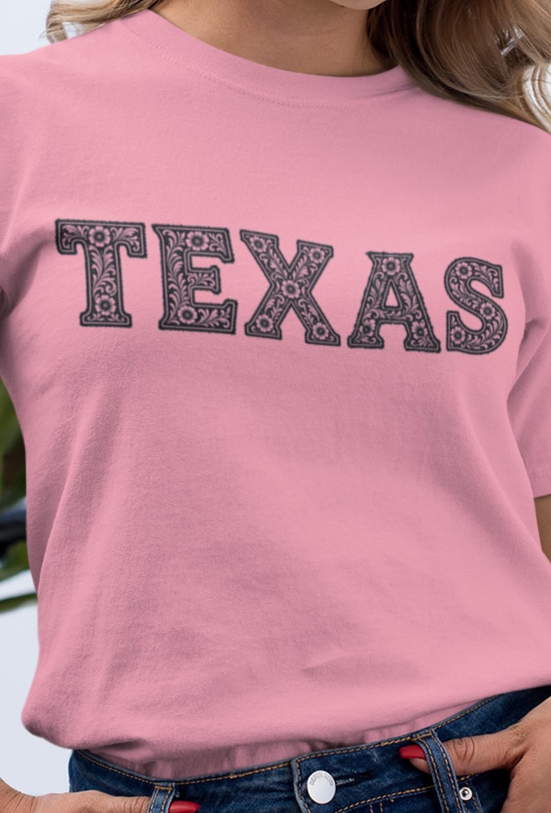May include: A light pink t-shirt with the word "TEXAS" printed across the chest in a decorative, floral-filled font. The letters are dark gray, and the shirt has short sleeves.