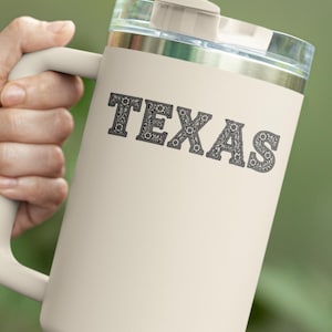 May include: Cream-colored travel mug with a handle and a clear lid. The word "TEXAS" is printed in a decorative font on the front of the mug. A green straw extends from the lid. The mug is held by a person.