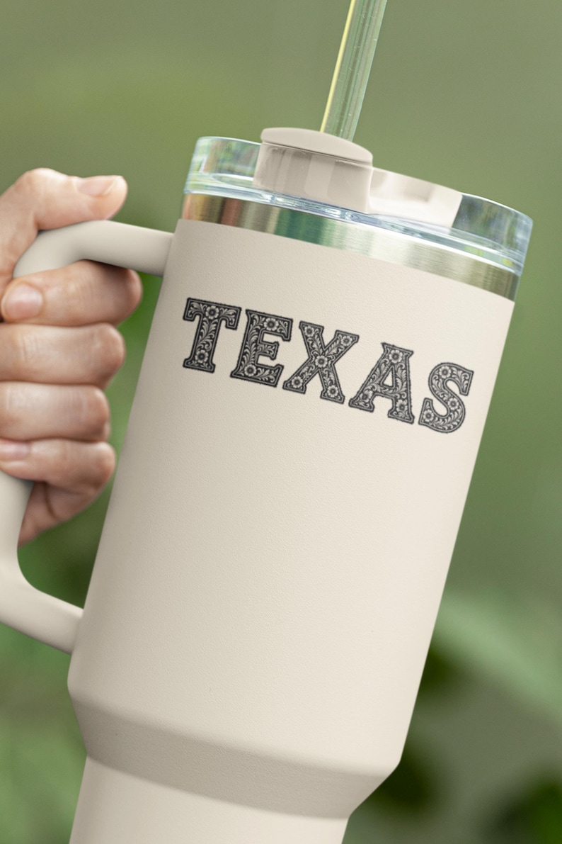 May include: Cream-colored travel mug with a handle and a clear lid. The word "TEXAS" is printed in a decorative font on the front of the mug. A green straw extends from the lid. The mug is held by a person.