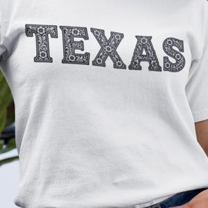 May include: White t-shirt with the word "TEXAS" in a decorative, floral-patterned font. The text is in a dark gray color, and the shirt is paired with blue jeans.