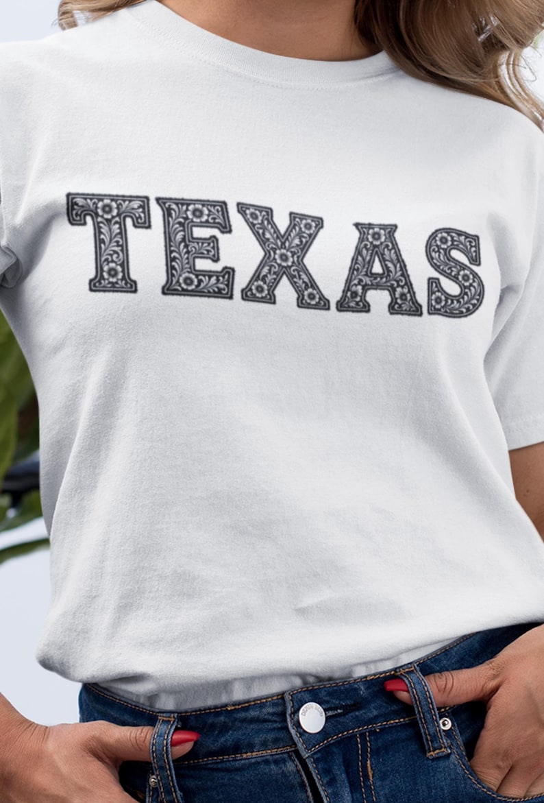 May include: White t-shirt with the word "TEXAS" in a decorative, floral-patterned font. The text is in a dark gray color, and the shirt is paired with blue jeans.